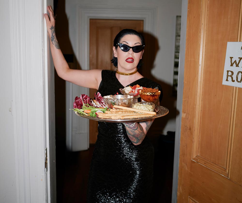 Shannon Martinez entering a room wearing a glamourous dress and sunglasses, carrying a platter of food.