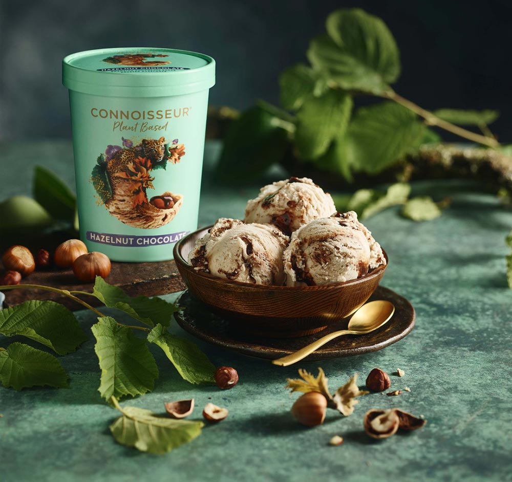 A tub of Connoisseur plant-based Hazelnut Chocolate ice cream sits on a dark green bench, surrounded by greenery and whole hazelnuts. In the foreground, three scoops of ice cream sit in a chocolate brown dessert bowl served with a gold spoon. 