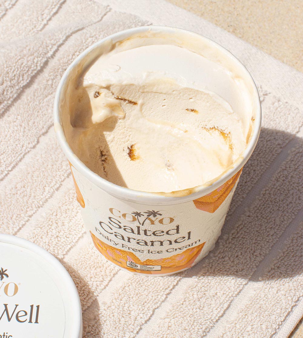 An overhead view of an open tub of COYO Salted Caramel ice cream on a beach towel, with scoops taken out, revealing the caramel swirls throughout. 