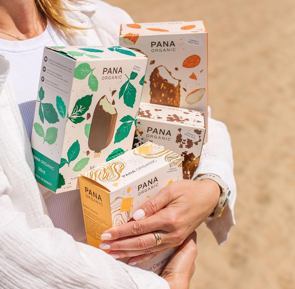 A person at the beach has their hands full with four different flavoured boxes of Pana Organic ice cream sticks.