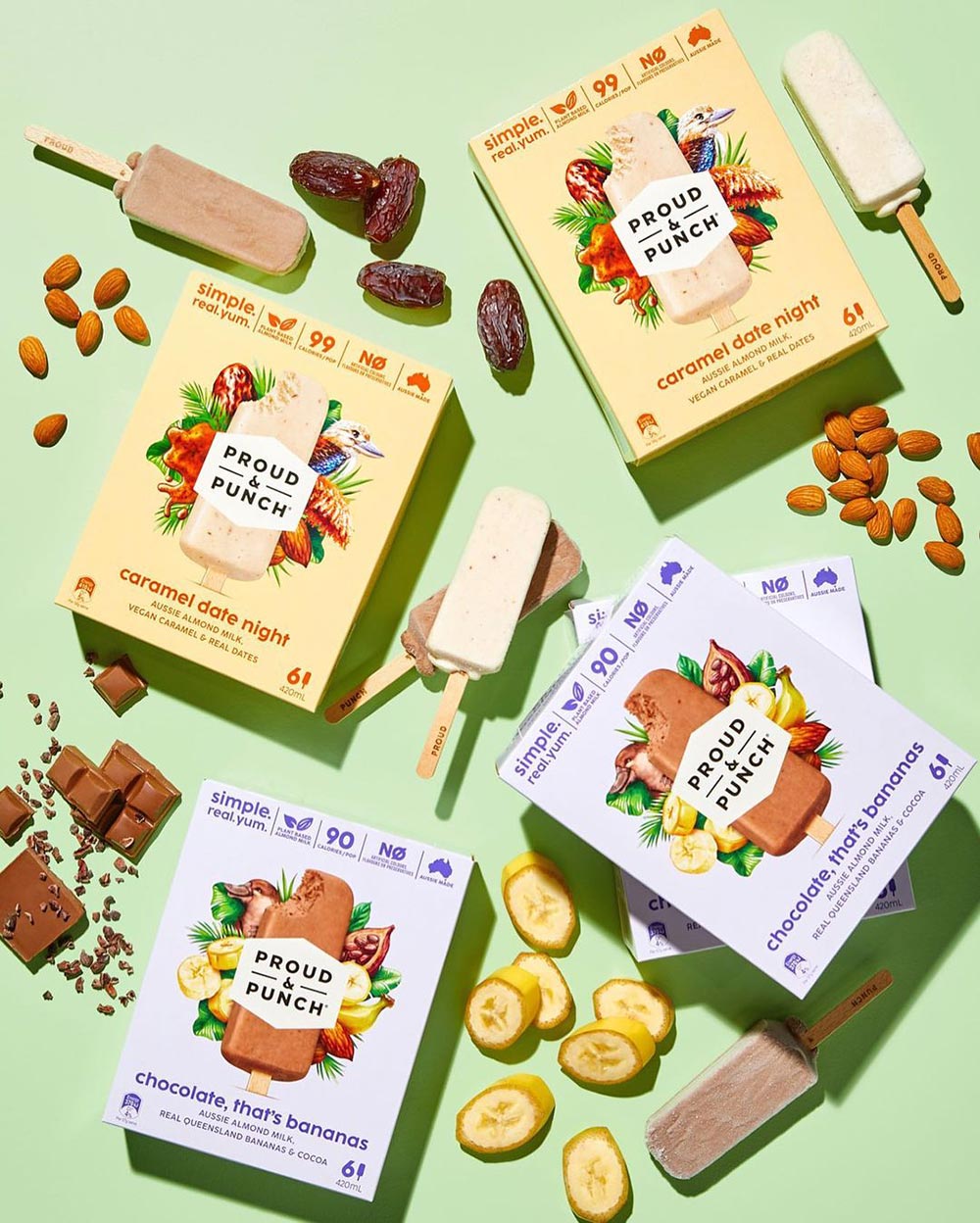 The packaging of four different flavours of Proud & Punch pops sit on a light green background, each with an open pop next to them in varying colours and surrounded by their respective natural ingredients. 