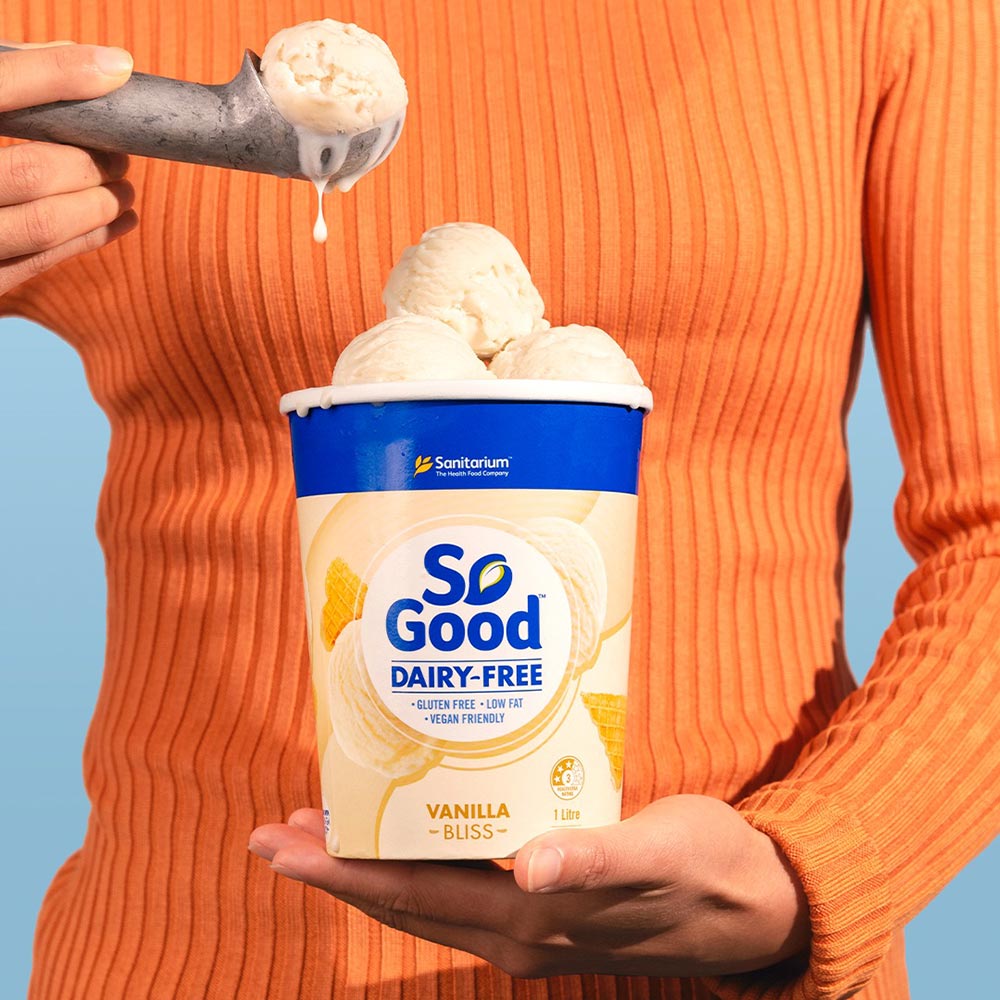 The mid-section of a woman in a bright orange top, holding a sub of dairy-free So Good ice cream to camera. In one hand, she holds a scoop, while the tub is overflowing with more creamy scoops. 