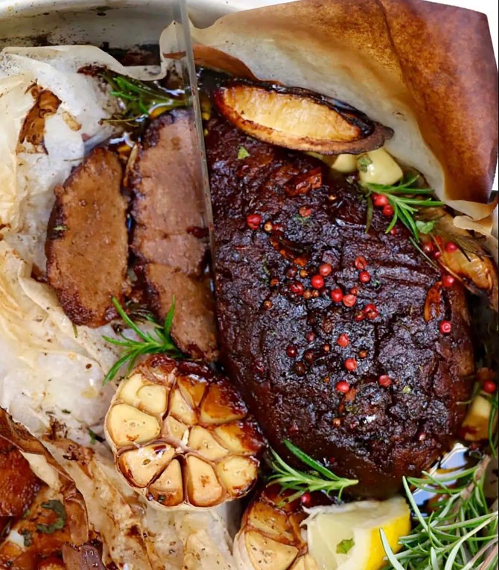 A juicy roast seitan 'lamb' is in a baking dish with lemon wedges and garlic.