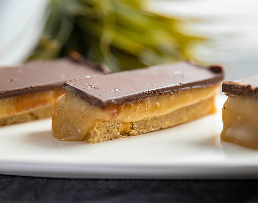 A close-up of a slice of gooey peanut butterscotch slice, with a biscuit base and topped with chocolate.