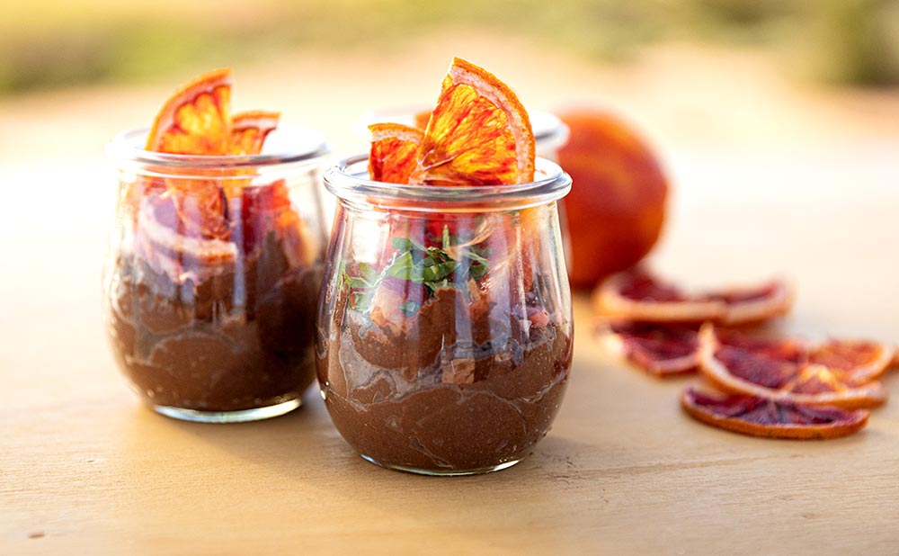 Two glass tumblers of chocolate mousse sit on a bench outside. They're topped with candied oranges.