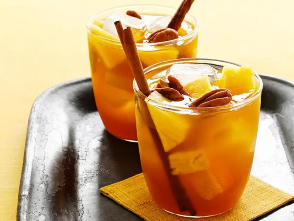 Two tumblers of cinnamon and pineapple punch sit on a tray. Straws have been fashioned out of cinnamon sticks and the drinks are topped with pecans.