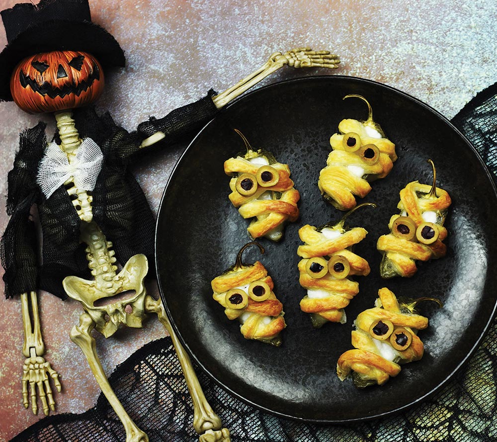 A bowl of stuffed jalapenos bandaged in puff pastry are decorated as mummies. A pumpkin skeleton prop sits next to the bowl. 