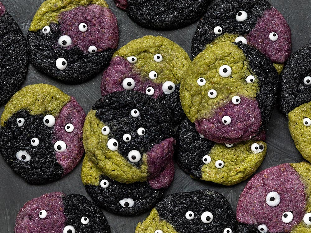 Cookies of green, black and purple with many edible eyes sit on a black tablecloth.