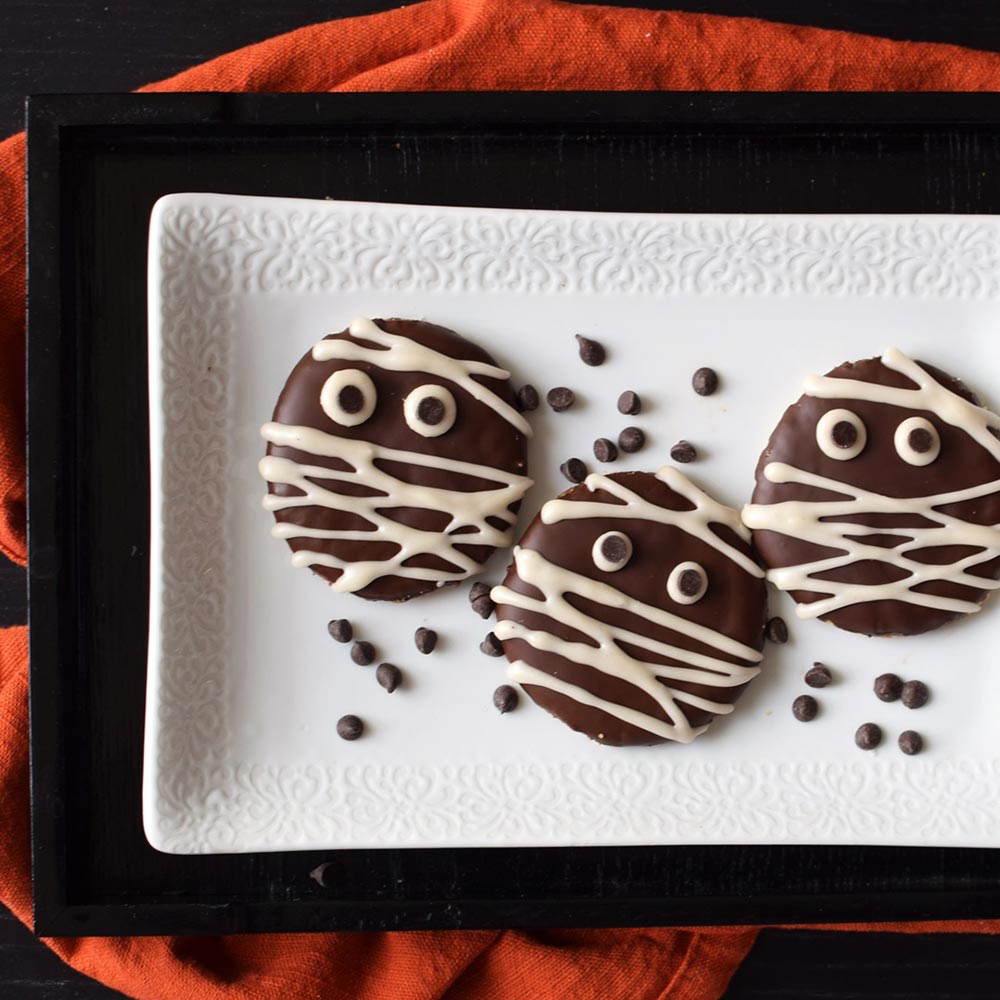 Round chocolate-coated biscuits on a white tray, with white chocolate drizzle acting as a mummy's bandage.