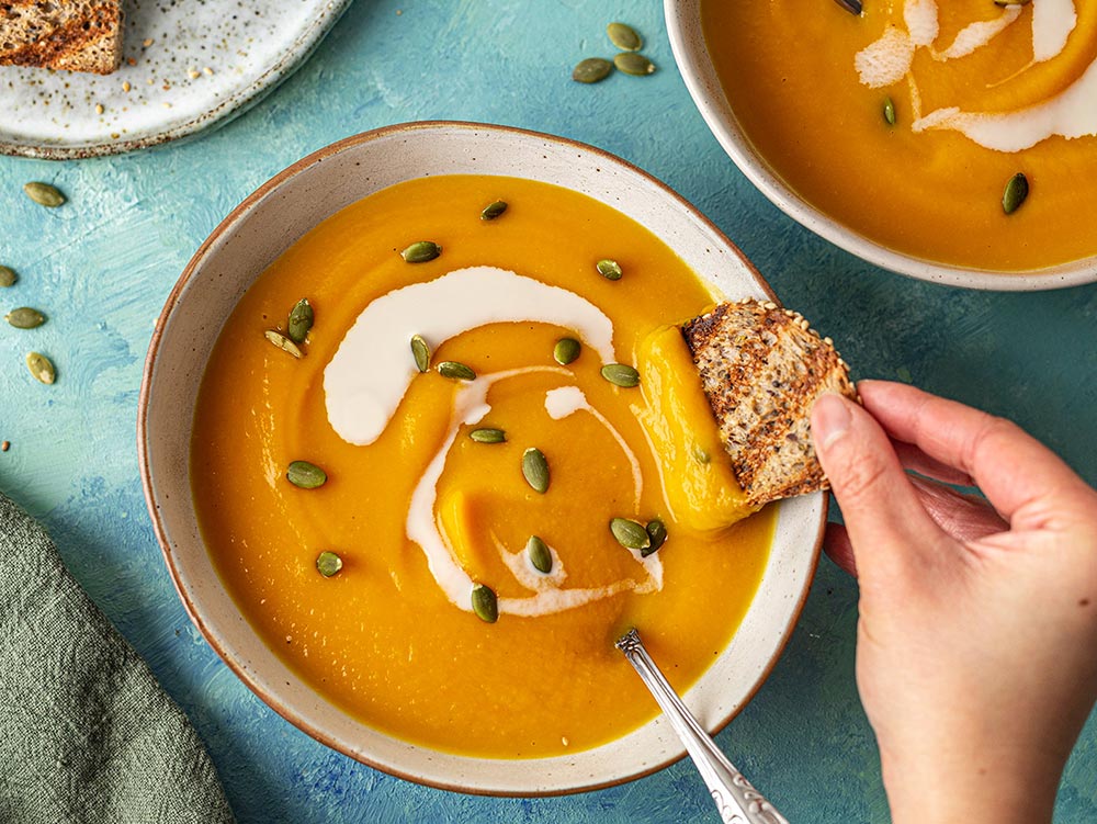 A hand reaches into a bowl of pumpkin soup to dip a slice of toasted bread.