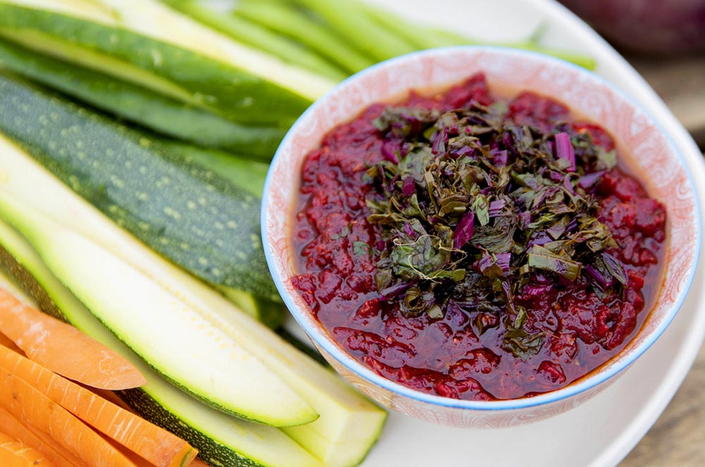 A deep red beetroot harissa sits in a small bowl with fresh vegetables for dipping off to the side.