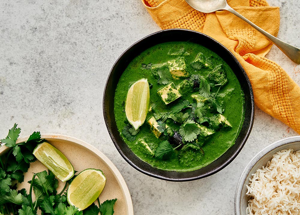 A vibrant forest green curry in a black bowl sits on a white benchtop.