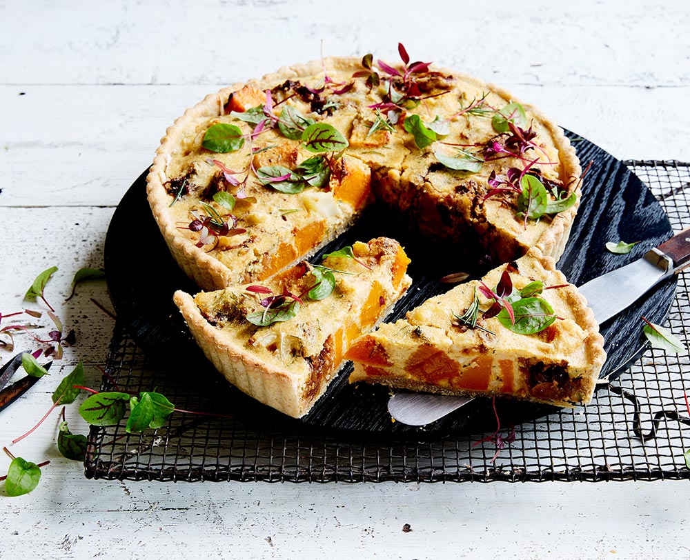 A whole tart sits on a cooling rack, with slices cut to reveal a filling of pumpkin and caramelised onion. 