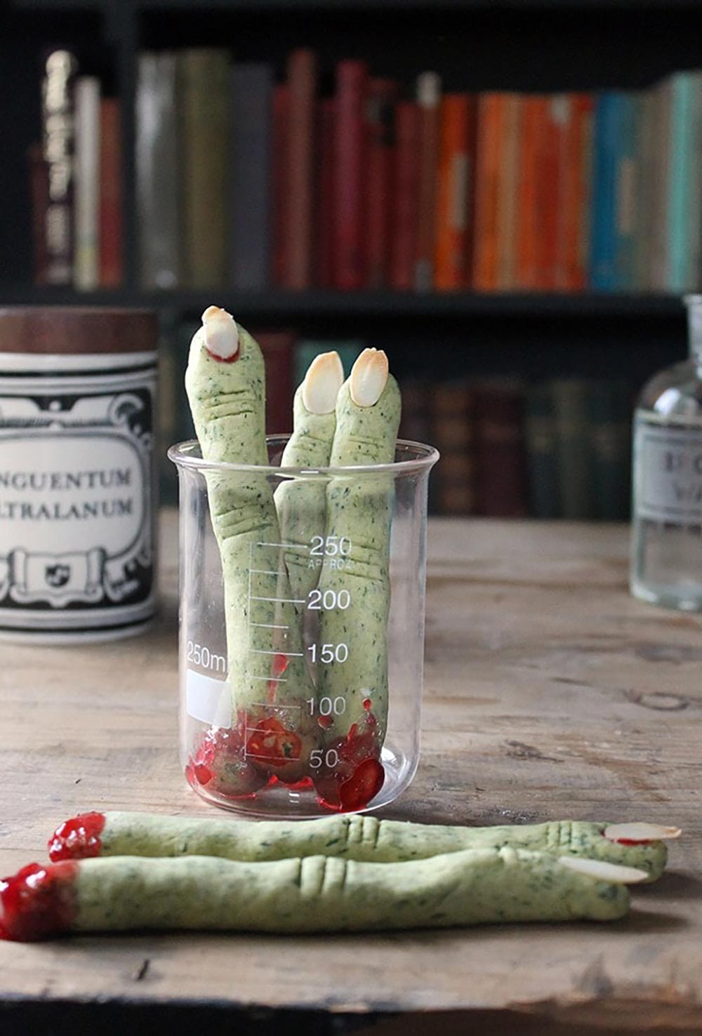 Green cookies in the shape of fingers sit in a beaker in an apothecary set up. They have a bloodied jam at the end on the knuckles.