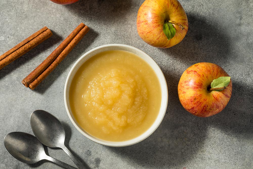 Overhead view of apple sauce in a ramekin, with full apples and cinnamon sticks laid to the side.