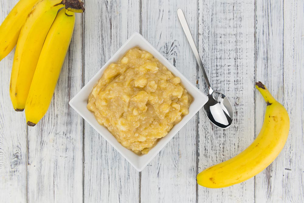 Mashed banana in a square bowl on a table. Full bananas sit to each side.