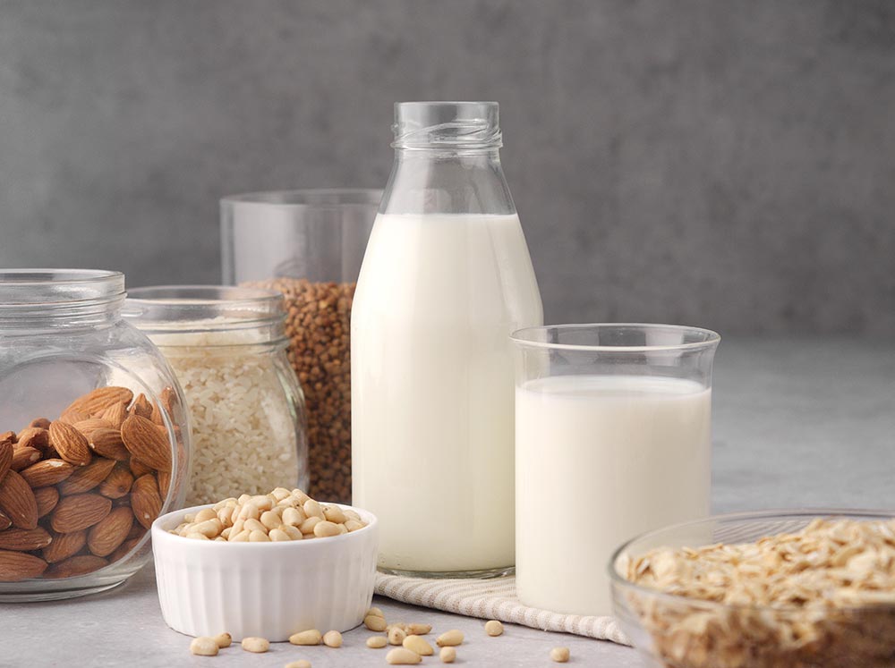 A plain glass jar of plant-based milk next to a full glass. Various nuts are in clear glass jars around them. 