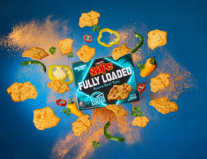 A promo pic of Arnott's Shapes, Fully Loaded Bueno Beef Taco flavour, on a blue background, with spice ingredients exploding out of the pack.