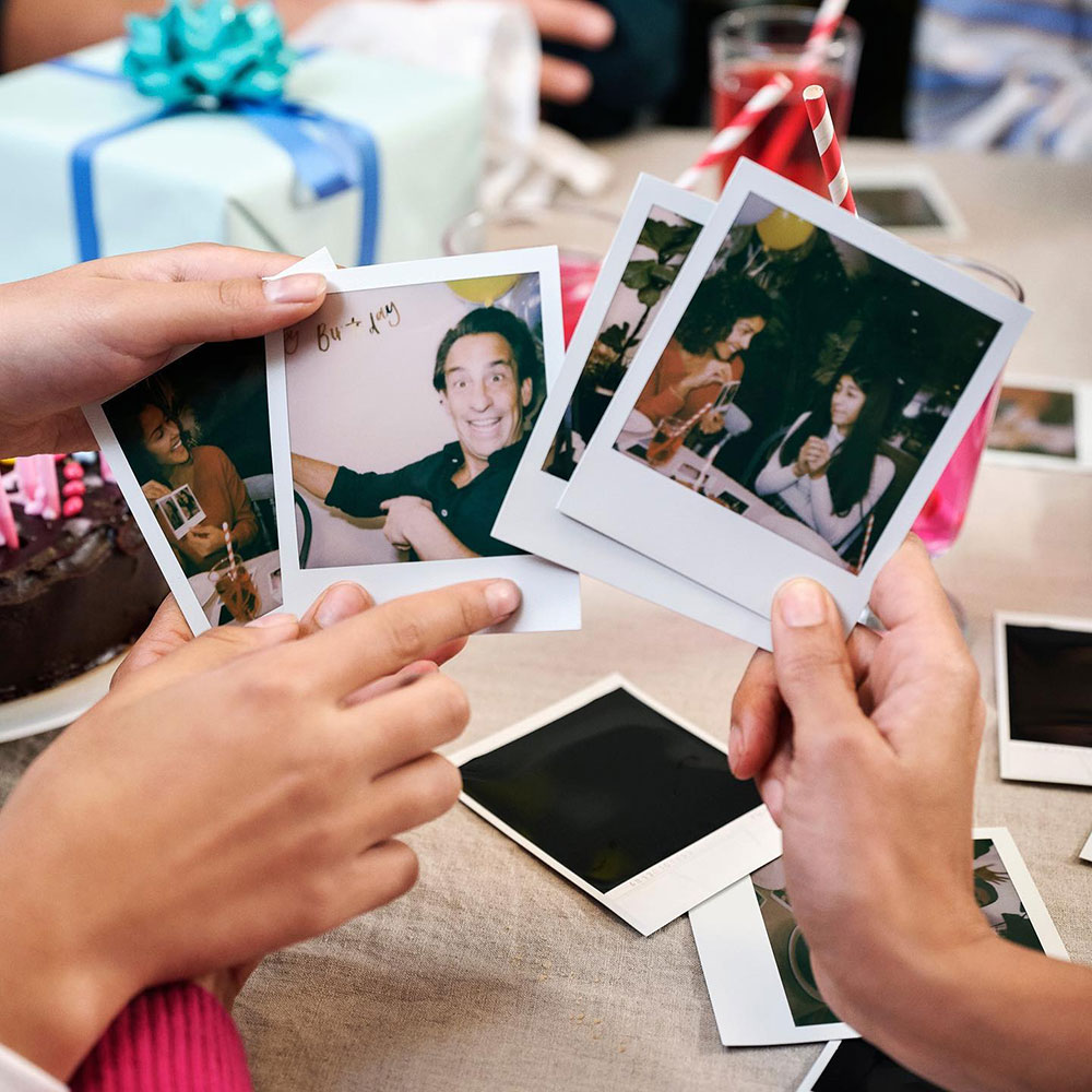 Hands hold up some polaroid photos featuring people at celebrations where food is involved. In the background, you can see drinks on a table with a wrapped gift. 