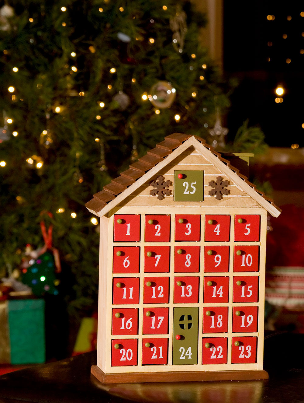 A beautifully crafted wooden house advent calendar sits in front of a Christmas tree.